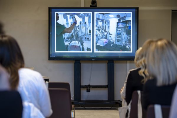Publika gleda prezentaciju na velikom zaslonu s fotografijama bolničke opreme i robota za kirurške zahvate.