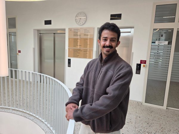 Smiling man in a brown fleece jacket stands by a white railing in a modern lobby with elevators and a wall clock.