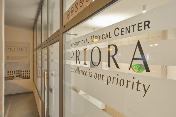Glass entrance of PRIORA International Medical Center with the slogan “Excellence is our priority” and clinic interior visible inside