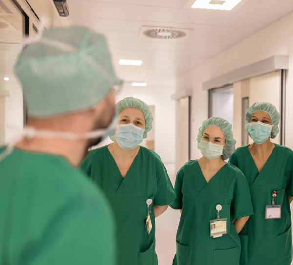 Tri medicinske djelatnice u zelenim odorama i maskama stoje u hodniku bolnice, slušajući kolegu u prvom planu.