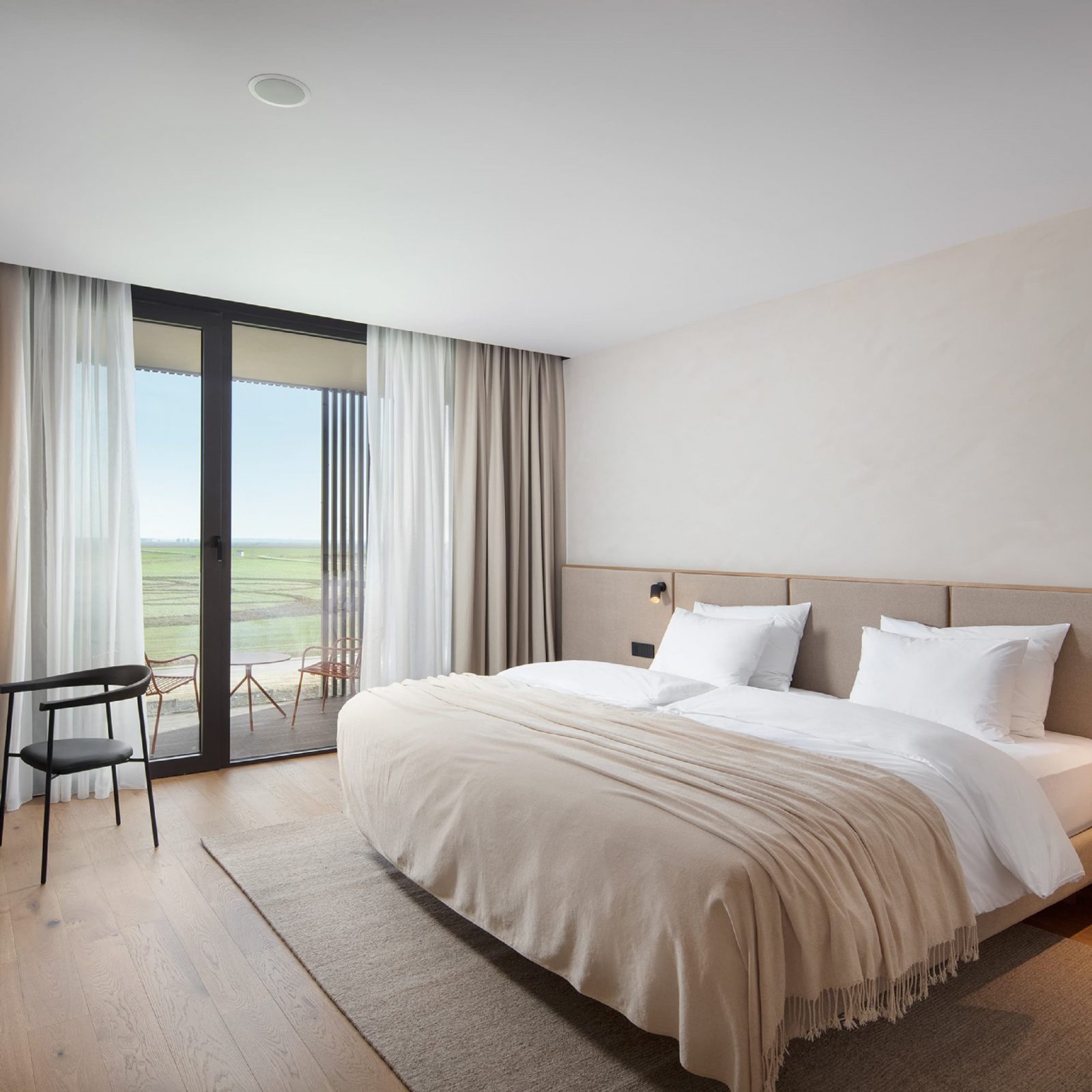 Moderna hotelska soba s velikim bračnim krevetom, svijetlim zavjesama i kliznim staklenim vratima koja vode na balkon s pogledom na prirodu.