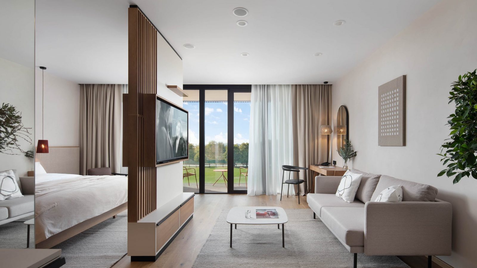 Elegant hotelska soba s modernim namještajem, uključujući krevet, kauč, televizor na zidu, radni stol i staklena vrata koja vode na balkon s pogledom na zelenilo.