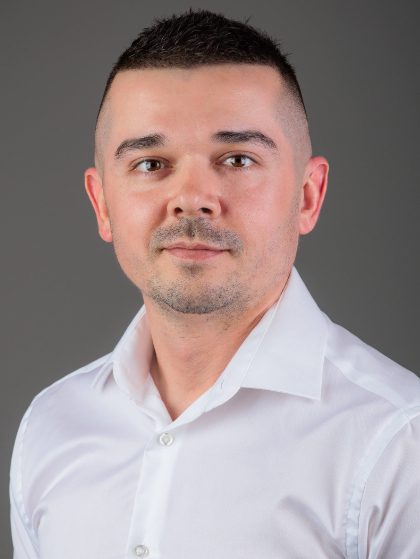 Muškarac s kratkom tamnom kosom u bijeloj košulji na sivom pozadinskom zidu.