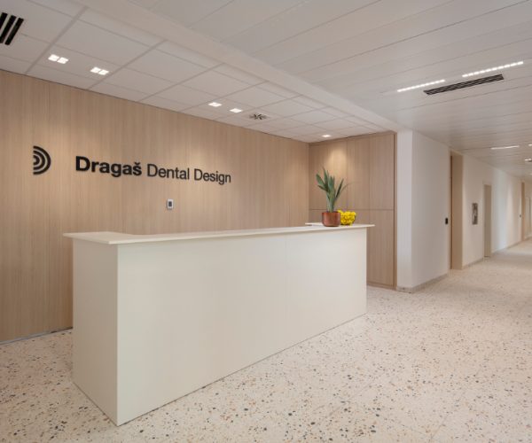 Recepcija u Dragaš Dental Designu s bijelim pultom, drvenim zidovima i ukrasnim biljkama te zdjelom voća na pultu.
