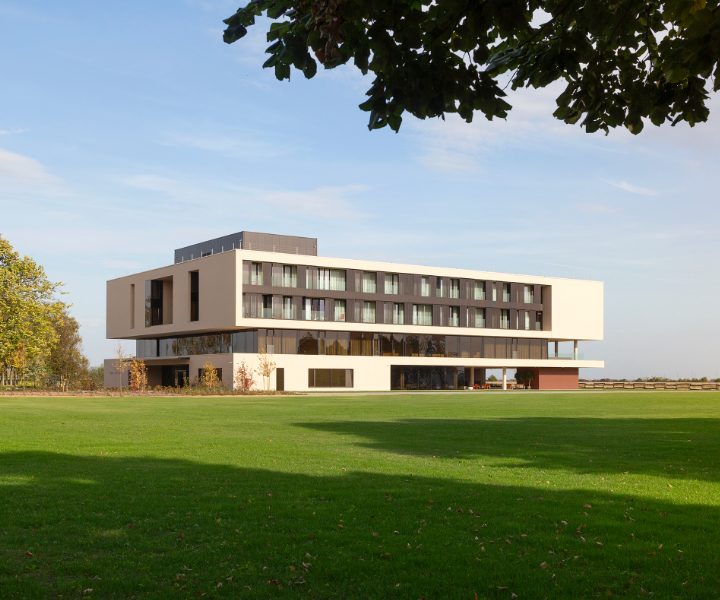 Moderna zgrada s ravnim krovom i velikim prozorima smještena na prostranom travnjaku, s drvećem u pozadini i lišćem u prvom planu, pod vedrim nebom.
