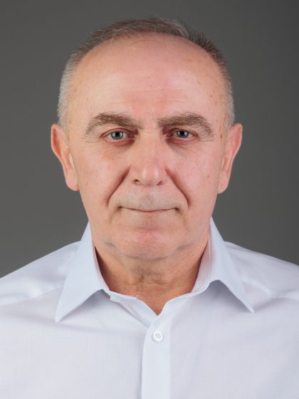 Muškarac srednjih godina u bijeloj košulji gleda ravno u kameru s blagim izrazom lica, na neutralnoj sivoj pozadini.