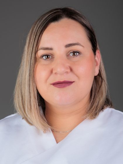 Žena sa svijetlosmeđom kosom i svijetlom bluzom, pozira za portret na sivoj pozadini.