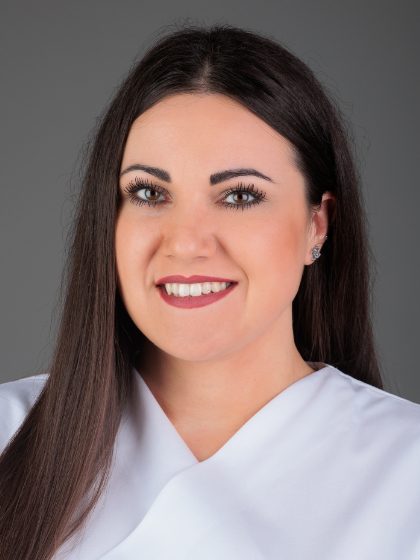 Žena s dugom tamnom kosom i bijelim odjevnim predmetom, nasmiješena, na jednobojnoj sivoj pozadini.