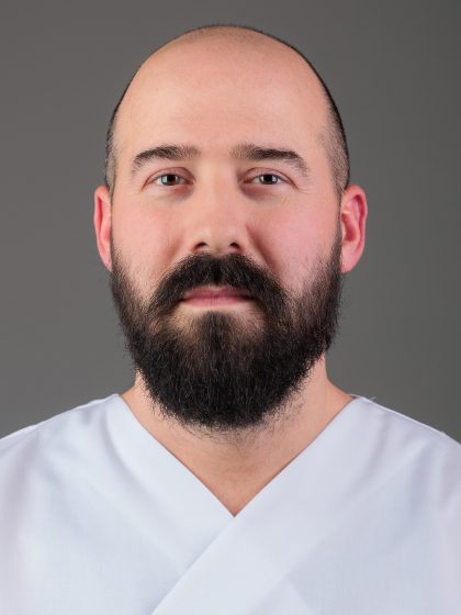 Muškarac s bradom i brkovima u bijeloj majici stoji ispred sive pozadine.