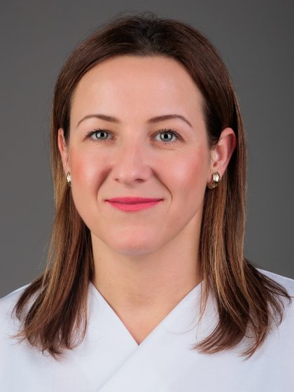 Žena smeđe kose, svijetlom puti, s naušnicama, odjevena u bijelu odjeću, pozira nasmiješena ispred sive pozadine.