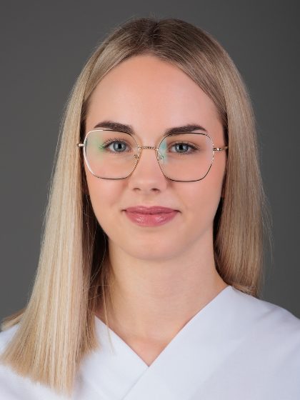 Žena s ravnom plavom kosom i zlatnim naočalama gleda prema kameri, obučena u bijelu odjeću.