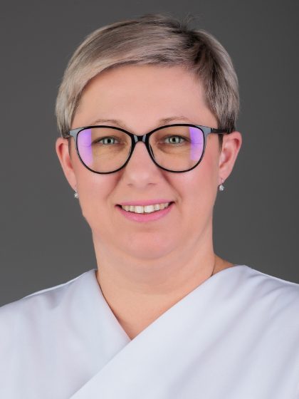 Žena sa svijetlom kratkom kosom i naočalama nosi bijelu odjeću i gleda prema kameri, na sivoj pozadini.