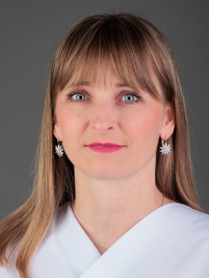 Žena s ravnom svijetlosmeđom kosom i plavim očima, nosi bijelu odjeću i srebrne naušnice s motivom zvijezde, na sivoj pozadini.