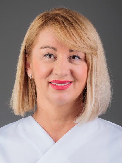 Žena s plavom kosom do ramena, obučena u bijelu košulju, nasmiješena i gledajući u kameru ispred sive pozadine.