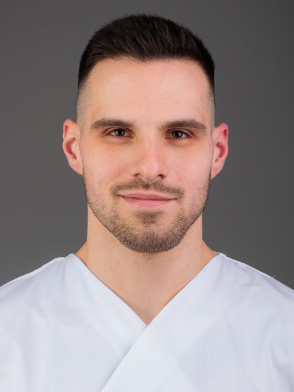 Mladić s kratkom smeđom kosom u bijeloj medicinskoj uniformi gleda ravno u kameru s blagim osmijehom, na sivoj pozadini.