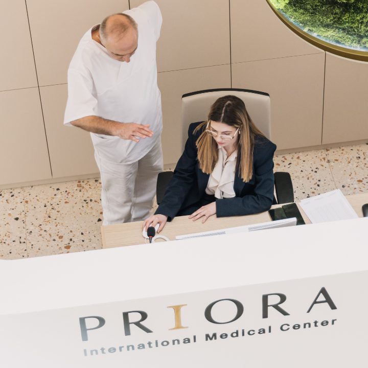Žena u poslovnom odijelu sjedi za stolom ispred konzole, dok muškarac u bijeloj odjeći pokazuje nešto na stolu u medicinskom centru PRIOIRA.