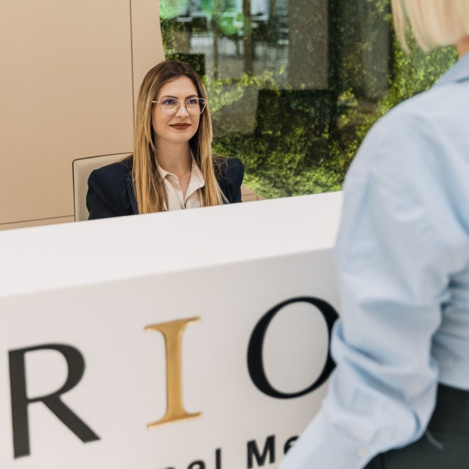 Žena nosi naočale i sjedi iza recepcijskog pulta, dok osoba stoji s druge strane. U pozadini se nalazi zid sa zelenim dekorativnim elementima.