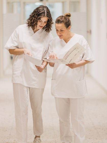 Dvije medicinske sestre u bijelim uniformama hodaju hodnikom i pregledavaju dokumente.