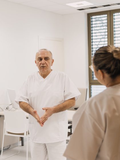 Liječnik u bijelom medicinskom ogrtaču razgovara s osobom u bež uniformi u modernoj bolničkoj sobi.