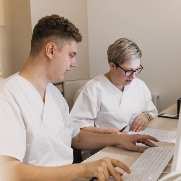 Dvoje osoba u bijelim medicinskim uniformama sjede za stolom i gledaju u računalski ekran, dok istovremeno vode bilješke na papiru.