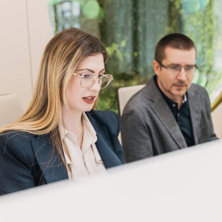 Dvoje ljudi sjede za stolom, fokusirani na rad na računalu, u modernom uredu s biljnim dekorom u pozadini.