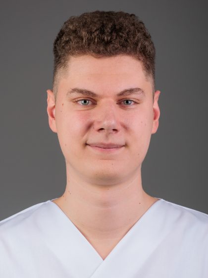 Mladić s kratkom kovrčavom kosom, obučen u bijelu majicu, pozira za portret na sivoj pozadini.