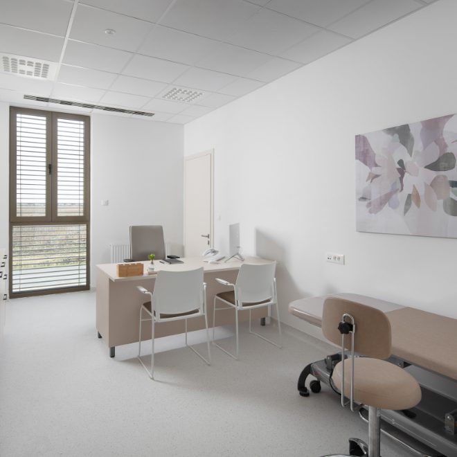 Uredno uređena liječnika ordinacija s radnim stolom, dvije bijele stolice ispred njega, krevetom za preglede sa strane i umjetničkim platnom na zidu.