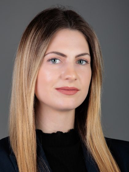 Žena s dugom, ravnom svijetlom kosom i tamnom odjećom pozira protiv jednostavne sive pozadine.