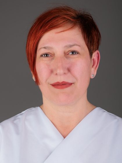 Žena s kratkom crvenom kosom nosi bijelu odjeću i naušnice, pozira na neutralnoj pozadini.