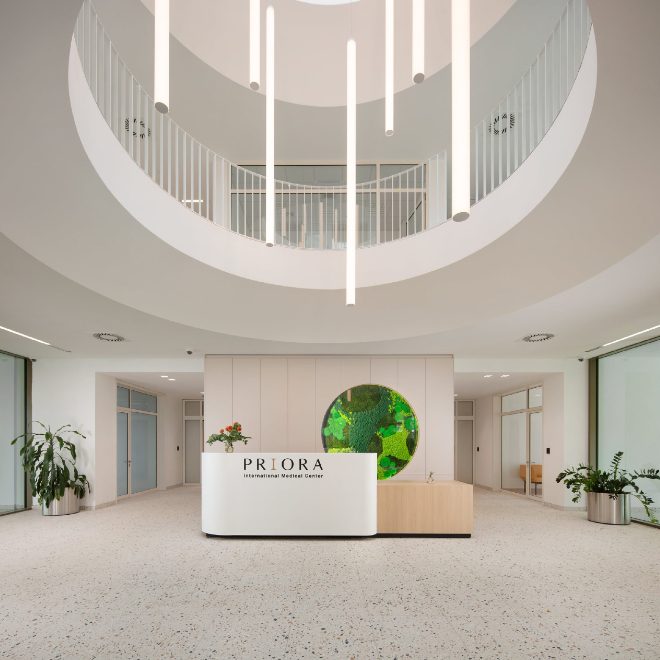 Unutarnji prostor moderne zgrade s recepcijom centra 'PRIORA' označenom na pultu, okružen biljkama i odiše minimalističkim dizajnom s visećim svjetiljkama i prostranim bijelim zidovima.