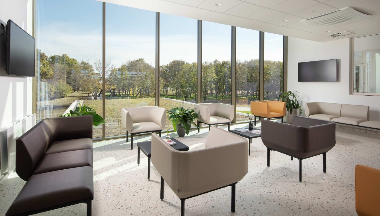 Svijetla čekaonica s modernim namještajem, uključujući smeđe i bež fotelje te sofe, i velikim prozorima koji pružaju pogled na zelenu vanjštinu.