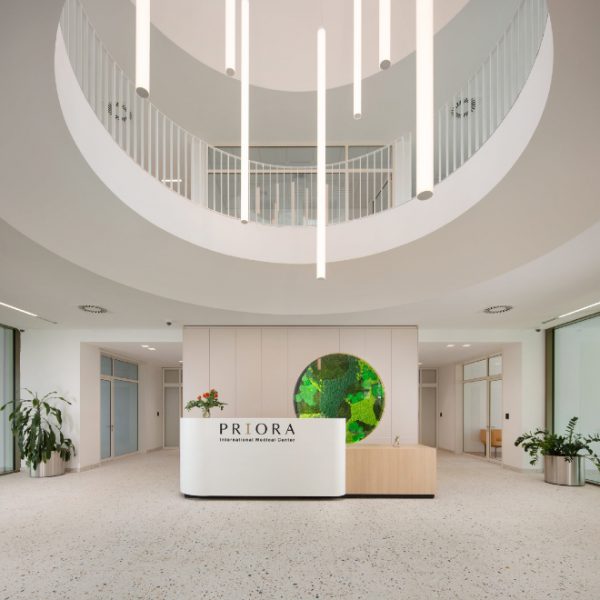 Moderno predvorje s bijelim recepcijskim pultom na kojem piše 'PRIORA International Medical Center', okruženo biljkama, s kružnim svjetiljkama koje vise sa stropa.