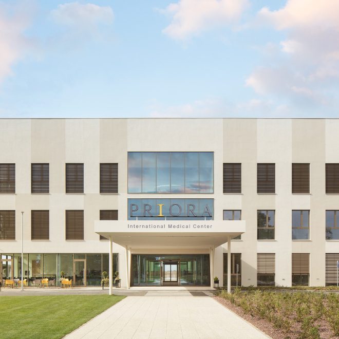 Modern medical center building with large windows and the sign 'PRIORA International Medical Center' above the entrance, surrounded by a manicured lawn and clear sky.