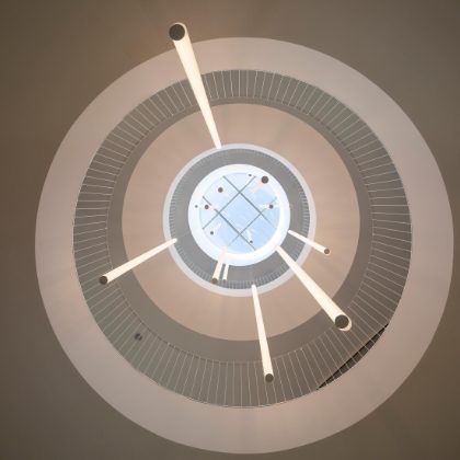 Pogled u modernu kružnu stropnu svjetiljku obješenu na visokom stropu, s višestrukim svjetlosnim zrakama koje se šire iz središta dizajna.