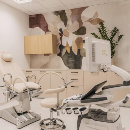 Interijer medicinske ordinacije s bež stolicom za preglede, ultrazvučnim aparatom i zidnom dekoracijom s cvjetnim uzorkom.