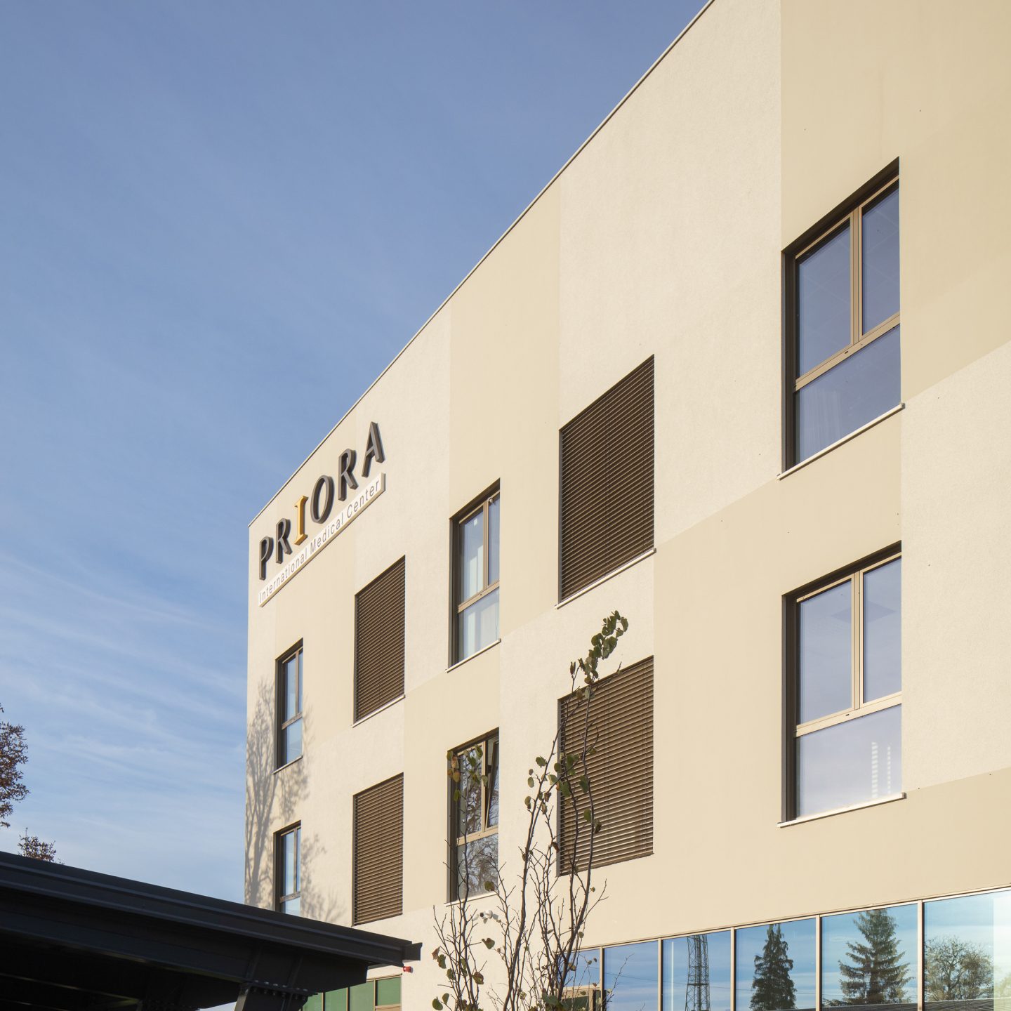 Zgrada s modernom fasadom i natpisom 'PRIORA Nonprofit Medical Center' na zidu, osvijetljena suncem i okružena prirodom.