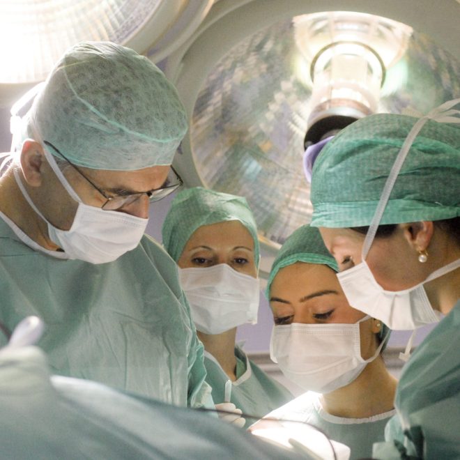 Kirurški tim obučen u zelene uniforme i zaštitne maske surađuje na operaciji pod snažnim svjetlom u operacijskoj sali.