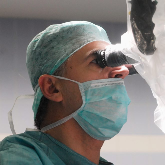 Kirurg nosi kiruršku masku i kapu dok koristi mikroskop tijekom operacije.