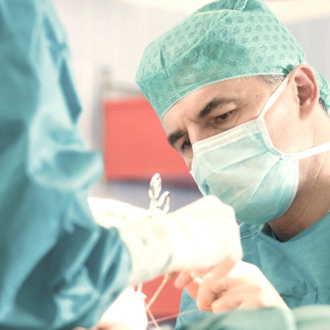 Kirurg u operacijskoj sali nosi zelenu kiruršku kapu, masku i rukavice dok fokusirano radi na pacijentu.