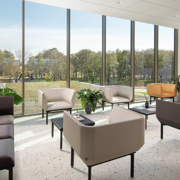Moderan uredski salon s udobnim foteljama i niskim stolovima, smješten uz velike staklene prozore s pogledom na zelenilo vani.