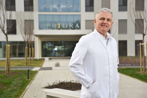 Muškarac u bijeloj liječničkoj kuti stoji ispred moderne zgrade s natpisom 'PRIORA'.