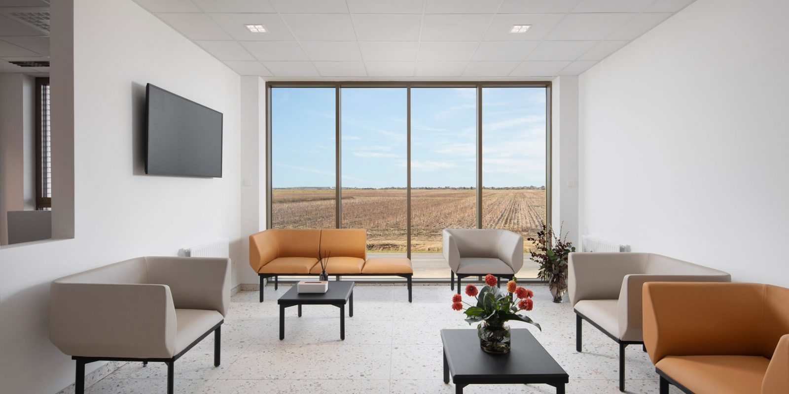 Moderna čekaonica s velikim prozorom koji gleda na polje, s krem i narančastim foteljama, crnim stolićem, televizorom na zidu i cvjetnim aranžmanom na stolu.
