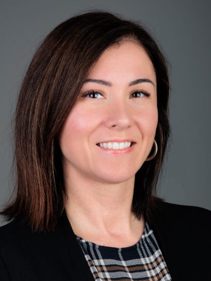 Žena s kratkom smeđom kosom, obučena u crnu jaknu i kariranu košulju, osmjehuje se na sivoj pozadini.