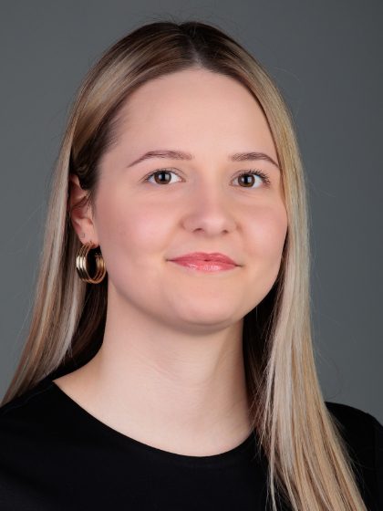 Žena sa svijetlom kosom nosi crnu majicu i zlatne okrugle naušnice, pozira za portret na sivoj pozadini.