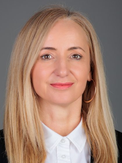 Žena s ravnom plavom kosom, nosi bijelu košulju i sako, s okruglim naušnicama, gleda prema kameri.