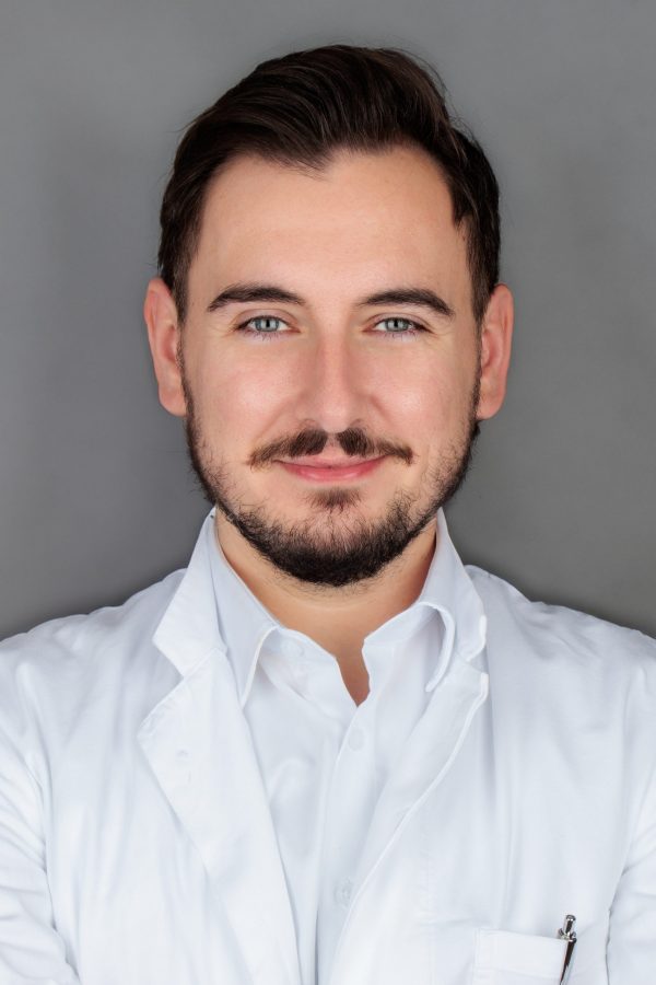 Mirko Dozan Muškarac tamne kose i brade u bijelom liječničkom ogrtaču nasmiješen je ispred sivog pozadinskog platna.