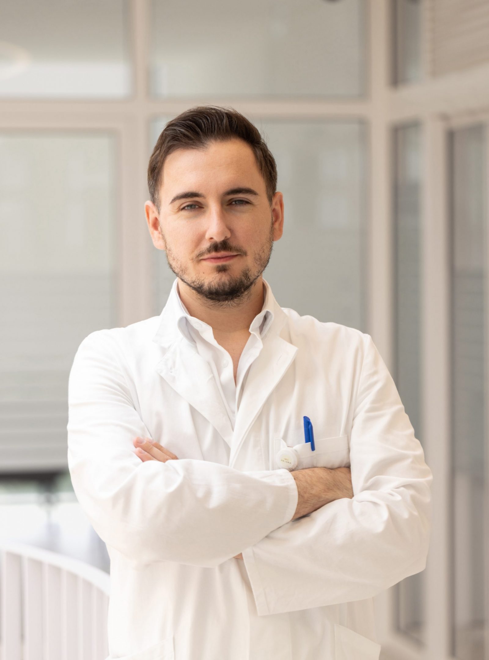 Muškarac u bijeloj liječničkoj kuti stoji s prekriženim rukama u hodniku, izgleda profesionalno i samouvjereno.