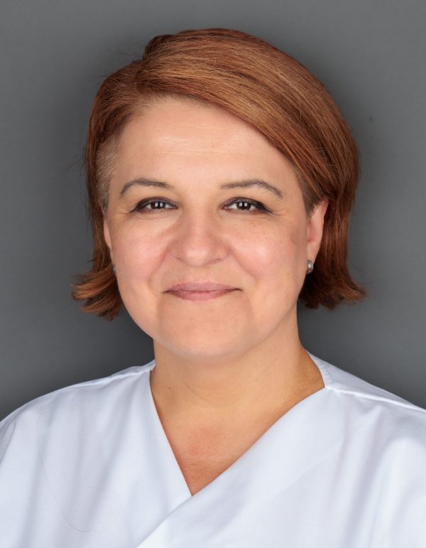 Žena smeđe kose u bijeloj uniformi pozira ispred sive pozadine.