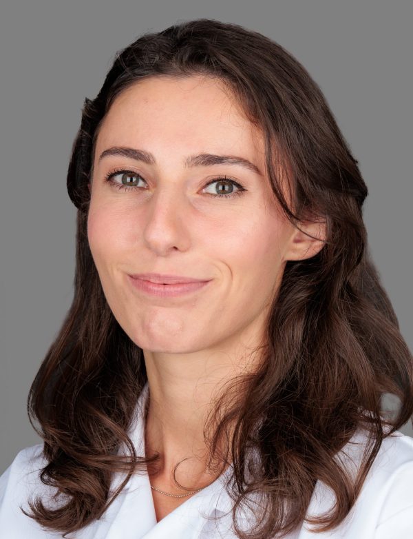 Žena s dugom smeđom kosom i bijelom odjećom gleda prema kameri, s blagim osmijehom na licu.