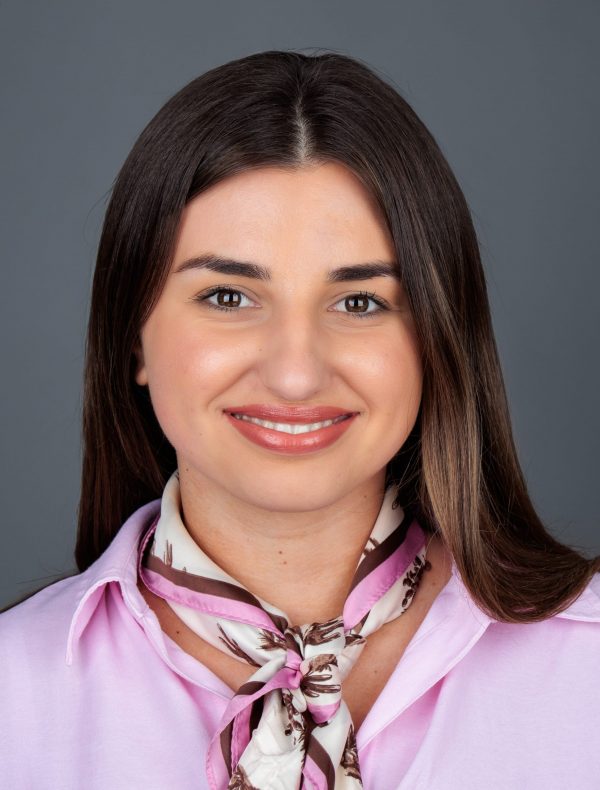 Žena s dugom smeđom kosom nosi ružičastu košulju i svijetli šal s uzorkom oko vrata, smiješi se prema kameri uz neutralnu pozadinu.